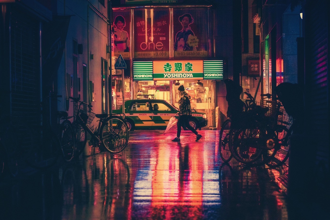 Parked bicycles line a narrow wet street illuminated by red, green, and amber signs at night.