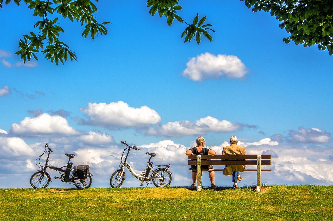 Two bicycles stand near a bench occupied by two people beneath a bright blue sky and scattered clouds.